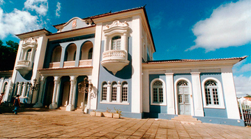 Foto em câmera baixa da fachada do Memorial de Medicina de Pernambuco. A foto contempla o céu azul em dia ensolarado e o prédio é pintado nas cores branca e azul.