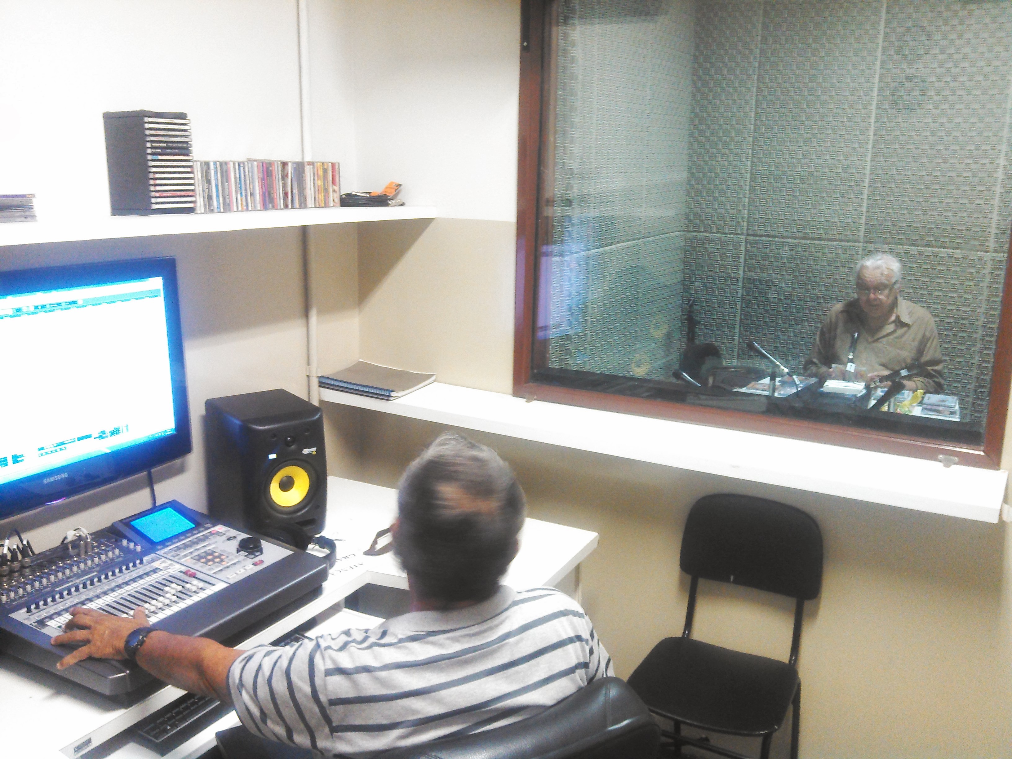 Técnico de som, operando equipamentos em uma sala, e professor apresentador gravando dentro do estúdio a locução do programa.